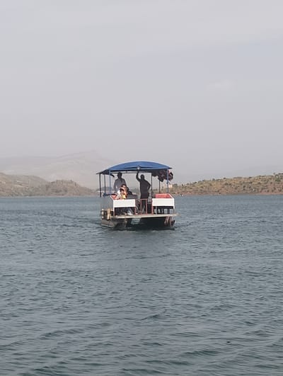 A medium shot captures a pontoon-style boat with a blue canopy navigating a large body of water, likely a lake or reservoir, in Bin El Ouidane, Morocco. Several people are visible on the boat; one person on the far right stands and waves their arm, while another person sits further back near the rear. At least two more individuals are partially visible towards the front of the boat, with one wearing an orange life jacket. The boat appears to be moving, leaving a slight wake behind it.
The sky is hazy and light grey, suggesting either an overcast day or atmospheric conditions reducing visibility. Distant, arid or sparsely vegetated hills and mountains frame the background, characteristic of the Moroccan landscape. The water is a deep blue-grey with gentle ripples across its surface. The overall impression is of a leisure activity, with people enjoying a boat ride in a scenic, rugged environment during daytime. No visible text is present in the image.