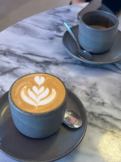 The image displays a close-up, high-angle view of two coffee servings positioned on a white marble-patterned table featuring dark gray veining. In the foreground, a gray ceramic cup, approximately half-filled, contains a light brown coffee beverage adorned with white foam latte art forming a stylized tulip or multiple heart design. This cup rests on a matching gray ceramic saucer, with a small silver spoon situated to its right. In the blurred background, a second, similar gray ceramic cup holds a darker brown liquid, likely an espresso, also placed on a matching gray saucer with a small silver spoon. Partially visible on the table in the background, near the second cup, is a smartphone and a segment of a person's hand. The specific location within Lordelo do Ouro e Massarelos, Portugal, is not discernible from the visual information provided.