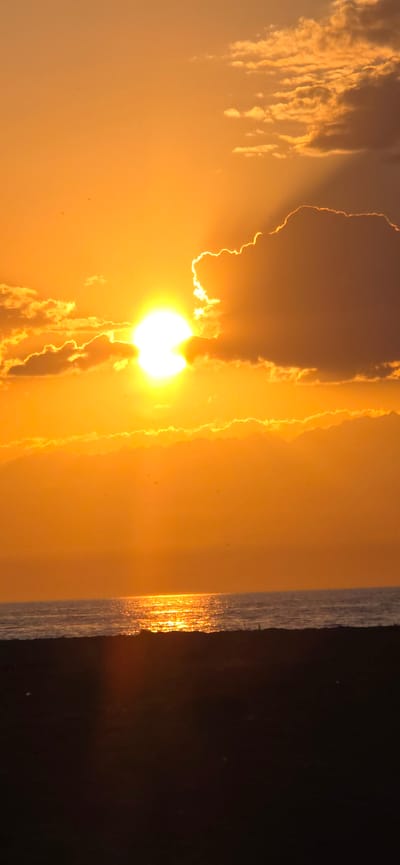 A vertical, medium shot captures a vivid sunset over the sea, with the sun partially obscured by clouds, casting a brilliant orange glow across the sky. The sea, reflecting the sunset's light, shimmers with a golden path. In the foreground, a dark silhouette of land, possibly a beach or dunes, lies in shadow. The scene appears to be a natural landscape, likely a coastal area, and there is no indication of the city of Boz Beshik, Kyrgyzstan. The image is devoid of people, objects, or any specific actions or interactions.