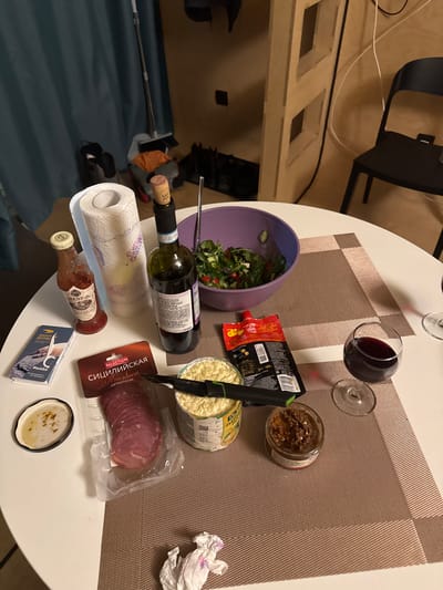 This image depicts an indoor scene in what is presumed to be a residence in Vasil'evo, Russia. A white, round table occupies the foreground, set with various food and drink items.
**On the table, from left to right:**
* A small glass bottle with a red liquid and a white label, possibly a sauce.
* A roll of white paper towels with a faint pattern.
* A dark green glass bottle of red wine, partially consumed, sealed with a cork.
* A purple plastic bowl containing a green salad composed of lettuce, cucumbers, tomatoes, and olives, with a metal serving spoon inserted.
* A vacuum-sealed clear plastic package of sliced red salami, labeled "Сицилийская КОЛБАСА" (Sicilian Sausage) in Cyrillic.
* An open metal can containing a chopped yellow food product, with a black utility knife resting horizontally across its top.
* A red squeezable pouch of food product with visible yellow granules on its packaging.
* A small glass jar containing a brown, chunky spread.
* A stemmed wine glass containing red wine, partially consumed.
* A second, empty stemmed wine glass is partially visible to the right.
* A small white dish with a dark rim holds a light-colored liquid topped with red flakes.
* A blue-grey booklet or card with text and an image, labeled "ROBOT ВОДНЫЙ" and "@sololab".
* A crumpled white paper or tissue is present on the lower right portion of the table.
**In the background:**
* Dark teal or blue curtains hang on the left.
* A floor mop with a grey handle leans against the wall near the curtains.
* Black footwear is visible on the floor below the mop.
* Light brown wooden wall panels or furniture extend across the center and right.
* White electrical cables run vertically along the light brown surface on the right.
* A black electrical socket is mounted on the light brown wall.
* A black, simple-designed chair is positioned on the right side of the frame.
No individuals are visible in the scene. The lighting appears to be artificial indoor illumination.