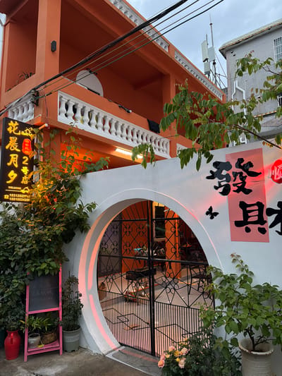 The image depicts the exterior of a business establishment, likely in the city of Longgang District, China, characterized by its vibrant orange facade. A prominent feature is a large, arched entryway with ornate black metal gates, leading into what appears to be a commercial space. To the left of the entrance, a black sign with yellow Chinese characters and the word "WELCOME" in English is illuminated, along with two phone numbers. Adjacent to the sign is a pink A-frame chalkboard displaying Chinese text, and below it are several potted plants. The white wall surrounding the archway is decorated with large black Chinese characters and a small black butterfly silhouette. Green foliage from nearby plants spills over the wall and frames the entrance. The building features a white balustrade on the upper level. Overhead, electrical wires crisscross the sky, and a cell tower is visible in the background. The lighting suggests it is either dusk or an overcast day, with warm light illuminating parts of the signage and the interior visible through the gate.