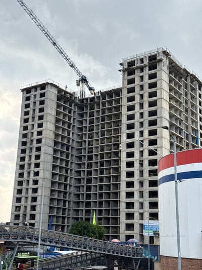 This image depicts a large-scale construction site located at Carrera 7 # 124-70 in Bogotá, Colombia. The primary subject is a multi-story concrete high-rise building, currently in an advanced stage of shell construction, featuring numerous empty rectangular window and door openings. A tall, black-framed tower crane extends diagonally over the building's top-left section, indicating ongoing work.
On the building's uppermost levels, several individuals, likely construction workers, are visible. One person stands on the highest left section, while a group is clustered near the crane's base on a lower, central roof section.
In the foreground, a metal pedestrian overpass with dark railings angles from the lower left towards the lower right. Several pedestrians are walking on this overpass; some individuals are visible beneath colorful umbrellas, suggesting informal vendors or protection from weather. Below the overpass, faint glimpses of what appears to be street-level activity and vehicles are present.
To the right, a prominent cylindrical structure, painted with a red top, a wide white band, and a central blue stripe, stands partially obscured. A street light pole is positioned in front of this structure. Further behind, a sign on a building advertises "APARTAMENTOS" (apartments), with visible text including "COY Y SIN SOFASA" and an address "cra 7 # 124-70," along with a price "DESDE $230 MILLONES." Green foliage from a tree is visible between the pedestrian overpass and the construction. The sky is uniformly overcast with gray clouds.