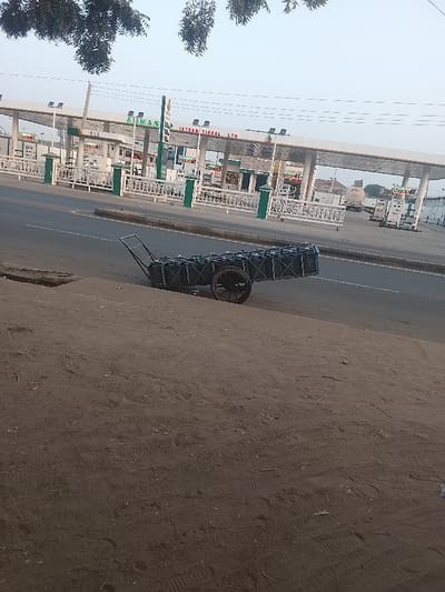 A two-wheeled cart, packed with black containers, rests on a curb alongside a road. In the background, a sprawling gas station complex with numerous pumps and signs is visible. The sky is overcast, and bare trees are in the top left corner of the frame. The foreground is dominated by a dirt or sandy surface, possibly a roadside verge.