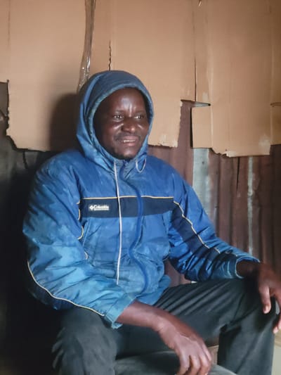 A medium shot captures a Black man in his late 30s or early 40s, sitting down and looking to his right, with a slight smile. He is wearing a blue hooded jacket with yellow trim and the "Columbia Sportswear Company" logo visible on the left chest. His skin appears to be dark, and he has short, dark hair. He is also wearing dark pants.

The background consists of rough, uneven walls made of what appears to be corrugated metal and cardboard. The lighting is dim, casting shadows on the walls and the man's face. The overall impression is of an indoor space that is sparsely furnished and somewhat dilapidated. The scene is likely a residential interior, possibly in Madalla, Nigeria, given the context.