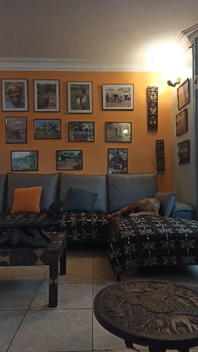 The scene depicts an interior living space in Kigali City, Rwanda. The dominant feature is an L-shaped sectional sofa with gray cushions, positioned against an orange-painted wall. A dark brown and white patterned throw blanket covers the seat and chaise sections of the sofa. A light brown dog is observed resting on the chaise section, with its head on a gray cushion. An orange throw pillow and a dark blue/gray throw pillow are placed on the main sofa section. Above the sofa, the orange wall features a gallery arrangement of twelve framed photographs, primarily depicting individuals, landscapes, and daily life scenes, potentially of African origin. To the right of the photo gallery, two elongated dark wooden decorative panels with carved faces and patterns are mounted vertically on the wall. An adjacent wall, lighter in color, displays several rectangular framed artworks. A wall-mounted light fixture illuminates the upper right corner of the orange wall. In the foreground, a dark wooden coffee table with geometric carvings stands in front of the main sofa. To the lower right, a small, round dark wooden side table, intricately carved with animal figures, is visible. The floor is tiled with light-colored, square tiles. The ceiling is white with visible crown molding.