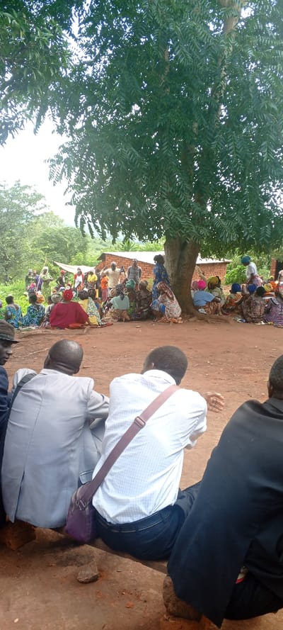 Under a large tree, a group of people are gathered in an outdoor setting. Most are seated on the ground, wearing colorful clothing, while others stand. The backdrop includes a building with a brick facade and surrounding green foliage. In the foreground, individuals dressed in formal attire, including suits and a shirt with a strap, are also seated, facing the larger group. The scene suggests a community meeting or gathering in a rural area.