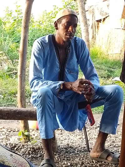 An adult male, appearing to be of African descent, is seated on a rustic wooden bench outdoors. He is wearing a light blue traditional outfit, consisting of a long-sleeved tunic and matching trousers. A light-colored skullcap covers his head. His gaze is directed downwards and slightly to his right. In his hands, he holds what appears to be a red object, possibly a phone case, and a pink object, which could be a phone. A second pink object is visible dangling from the bench to his right. The background consists of lush green vegetation and a structure made of woven material, suggesting a rural or semi-rural setting, potentially in Otulu, Nigeria. The ground beneath the bench is covered with small stones and gravel. The overall scene depicts a moment of casual repose.
