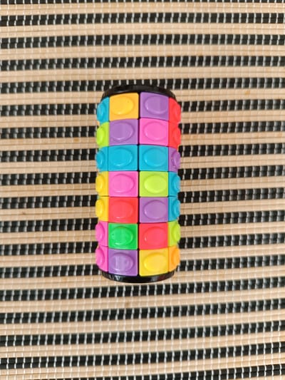 The image shows a cylindrical puzzle toy made of colorful interlocking blocks, placed on a striped placemat. The puzzle is cylindrical in shape and is composed of small, square blocks that are brightly colored in yellow, purple, cyan, pink, and green. The blocks are arranged in a way that suggests they can be rotated or manipulated. The toy is centrally positioned on a placemat with a distinct striped pattern of alternating light brown and black lines. The lighting suggests it is daytime, and the surface texture implies an indoor setting, possibly a table. There is no visible text in the image. The focus is entirely on the toy and the placemat, with no other objects or people present. The scene is simple and still, with the toy being the sole subject of interest. The colors of the puzzle are vibrant and playful, hinting at it being a children's toy. The composition is top-down, showcasing the entirety of the puzzle toy and a portion of the placemat.