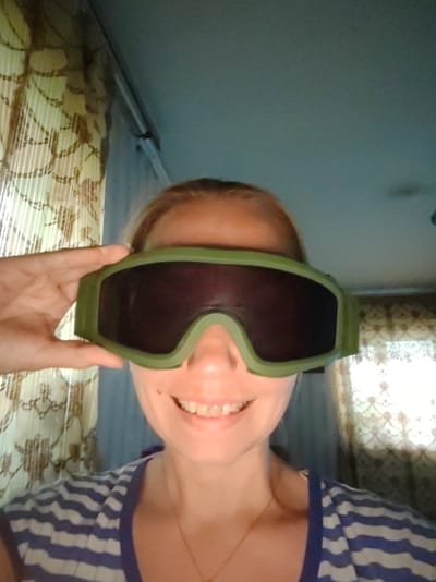 A close-up, front-facing selfie shows a person wearing large, olive-green goggles with dark lenses. Their right hand is raised to adjust the goggles on their forehead. The person has fair skin and a wide smile, revealing white teeth. They are wearing a blue and white striped t-shirt and a thin gold necklace. The background features a light blue ceiling and a patterned curtain with a floral or abstract design, illuminated by natural light, suggesting daytime. The scene appears to be indoors, possibly a home, with hints of a window and wall. The overall mood is lighthearted and playful. There is no visible text in the image. The location context is Чухлэм, Russia.