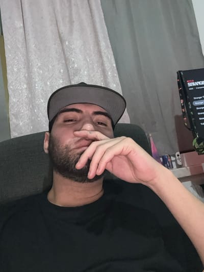 A man wearing a gray baseball cap and a black shirt reclines on a gray couch. His hand is near his face, with his fingers touching his beard and cheek. The background features a curtain with star patterns and a television screen displaying "Stranger Things" content. A Nintendo Switch is visible near the television.