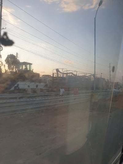A sunny road scene is viewed through a slightly dirty windshield. A street lamp and power lines run along the top left of the frame. Beyond these are several pieces of construction equipment, and a few smaller vehicles, along the shoulder of the road. A sandy-brown field extends out to the right. The sky is visible in the upper left.