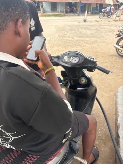 The scene is situated in Ikot Ekpene, Nigeria, depicting a roadside environment. In the foreground, a dark-skinned individual, viewed from the rear-right, is seated on a black scooter. This individual wears a black polo shirt with a white collar, black shorts, and a black slide sandal on the visible right foot. A red, yellow, and green beaded bracelet is worn on the right wrist. A black smartphone is held in the individual's right hand, with fingers touching the screen. The scooter's dashboard, displaying a speedometer and other gauges, is visible. A black hose extends downwards from the right side of the scooter, disappearing from view. Another dark-skinned individual, wearing a black T-shirt with a white graphic, is partially visible behind the primary subject's left shoulder.

The background consists of a light brown, unpaved road. Along the road, a row of single-story structures with corrugated metal roofs is present. In the far distance, two individuals are riding a motorcycle. The rear section of another motorcycle is partially visible on the far right of the frame. A white pole with a blue base stands vertically in the mid-ground.