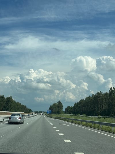 The image depicts a highway scene in the vicinity of Saue, Estonia, under a dynamic sky filled with cumulus clouds. A silver sedan with license plate "331 BTH" is traveling in the left lane of the multi-lane highway, foregrounded by the road surface marked with white dashed lines and rectangular reflectors. The highway is flanked by dense coniferous forests on both sides. Guardrails line the right side of the road, and a sound barrier is visible on the left. Several other vehicles, including trucks and cars, are observed further down the highway in the distance. The overall impression is a journey along a highway surrounded by natural landscape.