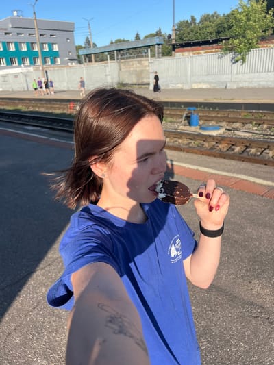 The media file depicts a young person enjoying an ice cream bar at a train station. The subject, with their hair in a messy bob, is shown from the chest up, wearing a blue t-shirt with a white graphic logo. They are holding the ice cream bar with their right hand, which has dark red nail polish. A black fitness tracker is visible on their left wrist. The person's face is partially obscured by a shadow from the sun, but their mouth is open as they lick the ice cream. A small star-shaped earring is visible in their left ear.
The background suggests an outdoor setting at a train station, with train tracks and a concrete platform visible. In the distance, there's a multi-story building with a teal and grey facade, and other people can be seen on the platform. The lighting indicates it's daytime, likely sunny. The overall mood appears casual and pleasant. The logo on the t-shirt is partially visible and appears to have text in Russian and a graphic element.