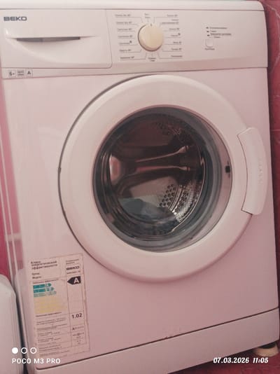 This image, taken on March 7, 2026, at 11:05 in Dmitrievskaya, Russia, displays a white BEKO front-loading washing machine, model WKN 60811 M, positioned in what appears to be a laundry area. The machine features a central beige rotary dial for program selection, with control labels predominantly in Russian indicating wash cycles like "Хлопок 90°" (Cotton 90°) and "Синтетика 40°" (Synthetics 40°). Specifications "6 kg," "800 rpm," and "A" (energy class) are printed on the upper left. The circular glass door of the drum is closed, revealing a dark item of clothing inside. An energy efficiency label, showing "Класс энергетической эффективности" (Energy Efficiency Class) with an 'A' rating, is affixed to the lower-left front panel. The washing machine is situated against a wall covered in reddish-brown tiles, with a vertical element, possibly a doorframe or another appliance, visible on the far left. No people or active interactions are present in the scene.
