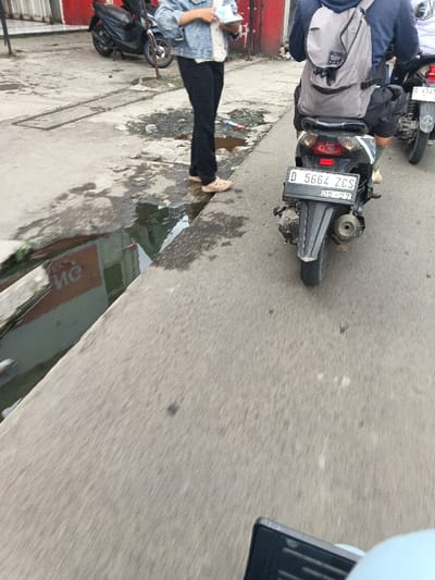 The image depicts a street scene in Margahayu, Indonesia. The foreground features an asphalt road, extending into the mid-ground. To the left, a concrete sidewalk runs parallel to the road, bordered by a narrow, water-filled gutter reflecting red and white architectural elements. Debris is present on the sidewalk and within the gutter.
On the sidewalk, a person is standing, visible from the mid-torso downwards, dressed in a light-colored denim jacket, dark trousers, and light brown clogs. This individual holds several white rectangular items in their hands. Behind them, a black scooter is parked next to a red-walled building with a closed metal storefront.
On the asphalt road, a black scooter, license plate D 5664 ZCS with a '09-27' validation date, is viewed from the rear. The scooter operator, wearing a dark hooded garment and dark shorts, carries a gray backpack. A second scooter, partially visible behind the first on the road, displays a partial license plate of D 432.... The environment represents an active urban thoroughfare.