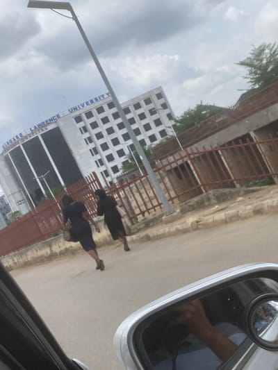 The image, taken from inside a vehicle, shows a view of Margaret Lawrence University in Kuchingoro, Nigeria. The university building is visible in the background, along with a sign bearing its name. Two pedestrians are walking on the sidewalk beside the building. The side mirror of the vehicle is visible in the foreground, reflecting an arm and part of the interior. The sky is overcast.