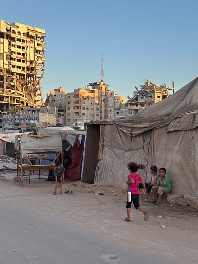 A group of children are gathered near a partially erected structure in Gaza. Three young boys, one in a green t-shirt and the others in white and dark shirts, are sitting and looking towards the camera. A girl in a pink t-shirt and dark pants is standing and appears to be in motion. She holds a white bottle in her left hand. Behind the children, a large, weathered tarp is stretched out, creating a makeshift shelter.

In the background, the scene depicts a devastated urban landscape. The remnants of bombed-out buildings stand as stark reminders of conflict. The architecture suggests a Middle Eastern setting, consistent with the provided location context of Gaza, Palestinian Territories. The sky is a clear, bright blue, indicating a sunny day. The lighting suggests it is either early morning or late afternoon.

The overall mood of the image is one of resilience and childhood amidst destruction. The children’s presence and apparent normalcy of their gathering contrast sharply with the surrounding devastation. The ground appears to be a rough, unpaved surface, likely a street or pathway. There is some debris scattered around. Some of the buildings have visible signage, but the text is too small and blurry to read. The activity captured is a moment of pause and social interaction among the children.