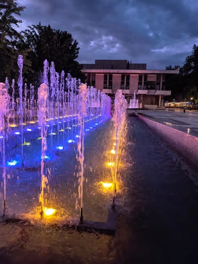 The image captures an outdoor urban scene at dusk or night, featuring a vibrant, illuminated water fountain system in the city of Svishtov, Bulgaria.

In the foreground and midground, a long, linear fountain runs from left to right, with numerous vertical water jets spraying upwards. The water jets are illuminated by colored lights embedded in the fountain basin. On the left side, the jets are predominantly purple/pink, transitioning through blue in the middle, and ending with yellow/orange on the right. The water flows into a narrow, dark channel alongside the main fountain.

In the background, positioned centrally, is a multi-story building with a flat roof, visible through and above the fountain jets. A sign on the building's facade, partially visible, reads "Народно читалище "Еленка и Кирил Д. Аврамови"" (National Community Centre "Elenka and Kiril D. Avramovi"), identifying the landmark in Svishtov. The building features multiple windows and a somewhat austere, likely concrete or stone, architectural style. To the left and right of the building, dark, dense trees are silhouetted against the twilight sky.

The sky above is overcast with dark, heavy clouds, indicating either a late evening hour or an approaching storm. Faint ambient light reflects off the clouds. To the far right, beyond the fountain and a paved plaza, an outdoor dining area with several white umbrellas and tables is visible, with indistinct human figures present in the distance. The ground immediately surrounding the fountain is a paved surface, part of a pedestrian area or public square. Artificial lighting from the fountain and distant streetlights is the primary light source in the scene.
