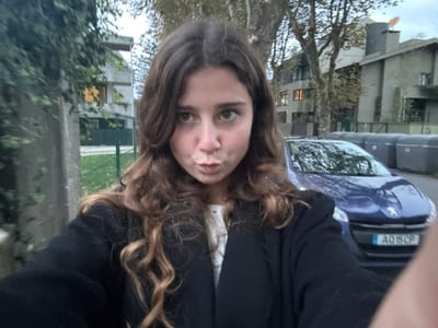 The image displays a close-up selfie of a young, fair-skinned female with long, wavy brown hair, looking directly at the camera with a pursed-lip expression. She wears a dark jacket over a light-colored top. The scene is a residential street in Aldoar, Foz do Douro e Nevogilde, Portugal.
In the background, a blue Peugeot vehicle with the license plate AQ16CP is parked on the street. To the right of the vehicle, multiple grey municipal waste bins are visible along the curb. Architectural structures include multi-story concrete buildings; one on the left features an illuminated window, while others further back are partially obscured by trees. A dense green vine covers a concrete wall on the left, bordering a grassy area with a green fence. Overhead, the sky is overcast, providing diffused natural light, and deciduous trees show sparse foliage.