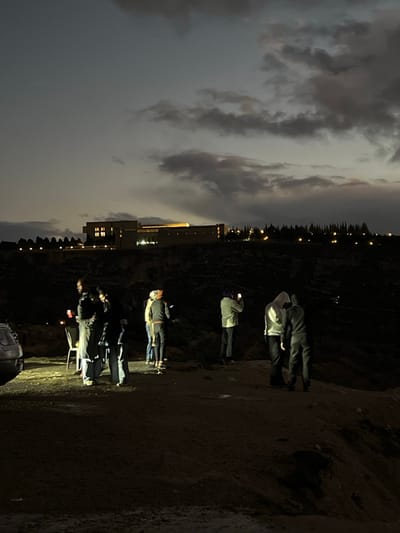 The image displays an outdoor nocturnal scene within Dummar Municipality, Syria. The foreground consists of an uneven, unpaved dirt or rocky terrain, serving as an elevated vantage point. Approximately eight individuals are present. On the far left, the rear section of a light-colored vehicle is partially visible, illuminated by a bright ground-level light source that also highlights the immediate area. Two individuals stand near this light, one possibly seated on a white folding chair to their left. Several individuals in the central group, including one wearing a light-colored hooded top, are observed holding and interacting with mobile phones, with some appearing to capture images or video. On the far right, two individuals, both wearing hooded garments, stand closely together.

In the background, across a dark, unlit valley or depression, a prominent multi-story building complex with numerous illuminated windows and sections is situated on a distant elevated plateau. A distinct line of dark, conifer-like trees and scattered additional lights are visible on the horizon behind and to the right of this building, indicating further development. The sky above is dark and heavily overcast with layered clouds.