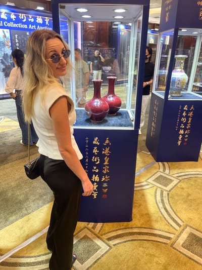 In the Futian District of China, a woman with blonde hair and sunglasses poses for a picture, leaning into the frame. She is wearing a white sleeveless top and black pants. The setting appears to be an indoor event, possibly an auction or exhibition, indicated by display cases filled with ornate vases. The walls are a deep blue, adorned with golden Chinese calligraphy, with some text visible including "al Collection Art Auction" and "藏藝術品拍賣". The floor is covered in a patterned yellow carpet. In the background, other individuals are visible, some looking at the displays, suggesting a public event. The lighting is bright, consistent with an indoor venue. The woman's pose and smile suggest a confident and positive demeanor.
