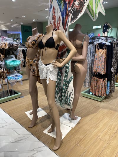 Three mannequins are displayed in a retail store. The mannequins are positioned to showcase swimwear. The mannequin in the front center is wearing a black bikini top and a white sarong skirt. To its left, another mannequin wears a gold and black patterned bikini. To the right, a third mannequin is partially obscured, but appears to be wearing a brown bikini. Behind the mannequins, racks of clothing are visible, including bikinis, dresses, and skirts. The floor is a light-colored wood laminate. A sign reading "LUXE SWIMSUIT" is visible in the background. The overall setting appears to be a department store or boutique specializing in swimwear. The location is identified as Wichit, Thailand.