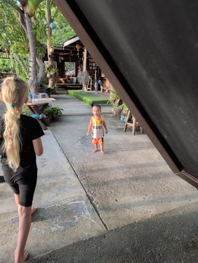 This media file depicts a scene in an outdoor setting, likely a residential area or a casual establishment in Ban Tai Subdistrict, Thailand. The orientation of the image is such that the subjects and environment appear to be sideways.

The main subjects are two people. In the foreground, a person with long blonde hair, wearing a black t-shirt, is seen from behind, with their arms resting on their lap and legs exposed. Their back is turned towards the viewer. In the middle ground, a young child is standing on a concrete surface. The child is wearing a yellow and white sleeveless shirt with orange shorts. They are looking towards the camera with a neutral expression.

The setting appears to be outdoors with a concrete walkway or ground. To the left of the child, there are lush green plants, trees, and some wooden structures that suggest an outdoor seating area or a garden. In the background, a fan is visible, along with what looks like a rustic, open-air building with various items displayed, possibly decorative objects or merchandise. Sunlight suggests it is daytime, with shadows cast on the ground, indicating it might be late afternoon. The overall atmosphere seems relaxed and casual.

There is some visible text on the child's shirt, which appears to be "BISTRO & HOME" in a stylized font.