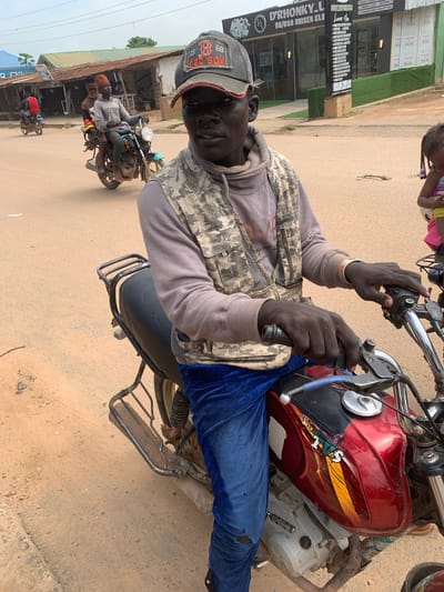 This media file, taken in Shagamu, Nigeria, captures a high-angle, portrait-oriented view of a man riding a motorcycle. The man is wearing a grey hoodie, a camouflage vest, blue jeans with a rip on the knee, and a grey baseball cap with "RED SOX" and numbers visible on it. He is looking directly at the camera with a neutral expression. His hands are on the handlebars, with one hand gripping a lever. A red and yellow sticker is present on the motorcycle's fuel tank, and a blue object, possibly a water bottle, is attached to the bike.
In the background, a young child with braided hair is partially visible at the top of the frame, wearing a pink and white striped top. To the left, a storefront with signage is visible. The text on the signage includes "D'RHONKY_L", "UK/USA UNISEX CLO", "SHOES", and phone numbers. The ground is a dirt or gravel road. Other motorcycles and people are visible further down the road, suggesting a busy street scene. The overall lighting suggests it is daytime, and the weather appears to be overcast. The image is oriented sideways.