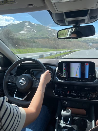 The image captures an interior view from the driver's perspective of a Nissan vehicle traveling on a road in Tsovagyugh, Armenia. The dashboard shows a map displaying the M-4 route. The driver's hand is on the steering wheel, and the gear shift is in park. The exterior shows a mountainous landscape with clear skies.