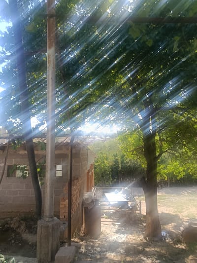 The image depicts a sun-dappled outdoor scene in a rural area, likely the village of Булак-Башы, Kyrgyzstan. Strong sunlight filters through the dense green foliage of trees, creating distinct beams of light that streak across the frame. In the foreground, a partially constructed brick building with a corrugated metal roof is visible. A weathered metal pole stands prominently to the left of the building, supported by a concrete base. To the right of the building, a large tree with a thick trunk stands in a yard. The ground appears to be a mixture of dirt and sparse vegetation. In the mid-ground, there is a collection of scrap metal and other discarded items piled on a metal frame, possibly a makeshift workspace or storage area. The overall impression is one of a humble, rustic setting under a bright, sunny sky.