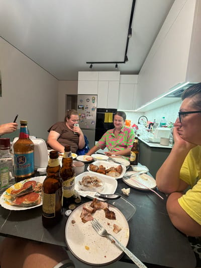 A group of women are gathered around a table in a kitchen, enjoying a meal and drinks. The table is laden with various dishes, including what appears to be grilled meats, fish, and pickled items, along with bottles of beer and what looks like vodka. The woman in the foreground, on the right, is wearing a yellow t-shirt and black-rimmed glasses, resting her chin on her hand. To her left, another woman in a bright pink and green patterned shirt is looking down at her phone. Further back, a woman in a brown t-shirt is also looking at her phone. The kitchen is modern with white cabinets and black countertops, illuminated by overhead lighting. The overall scene suggests a casual gathering or celebration. The image was taken in the city of Novorossiysk, Russia.