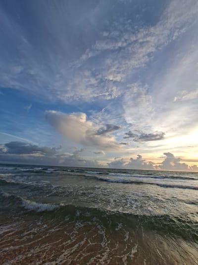 This is a sideways, close-up shot of the ocean and sky. The sky is filled with scattered clouds, some wispy and white, others darker and more dense. The sun is setting or rising, casting a warm, golden light on the horizon, which is visible as a thin line between the sea and sky. The clouds in the upper portion of the image are illuminated by this light, creating a beautiful gradient of blues, whites, and yellows.

The ocean takes up the right side of the frame. Small waves are breaking near the shore, creating white foam that washes onto the sandy beach. The water appears dark green and murky, with sunlight reflecting off its surface. The beach itself is a reddish-brown color, visible as the waves recede. The overall impression is one of a peaceful and serene coastal scene at dusk or dawn.

The image lacks any visible people or objects, focusing solely on the natural elements of the sky and sea. The location cues, such as the reddish sand and the specific way the waves are breaking, may suggest a tropical or subtropical beach. The time of day is clearly indicated by the dramatic lighting of the sky, and the weather appears to be fair with some clouds. The mood is tranquil and picturesque.