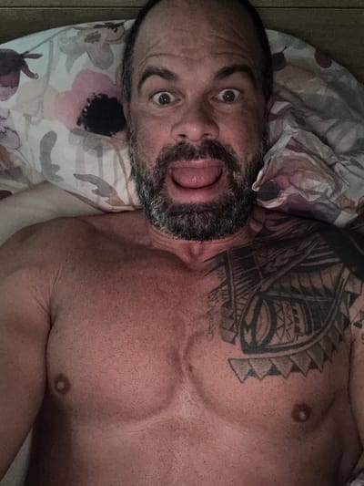 The image displays a shirtless man lying on a bed, viewed from a top-down perspective and rotated clockwise. The man has a beard with touches of grey, dark hair, and dark eyebrows. His eyes are wide open, and his mouth is agape, revealing his tongue, creating an expressive, possibly surprised or playful facial expression. He exhibits a muscular build, with clear definition in his chest and shoulders. A large, intricate dark tattoo with tribal or Polynesian-inspired patterns covers his left shoulder and extends onto his chest. His skin is fair, and some body hair or stubble is visible on his torso.
He is resting on a pillow adorned with a floral design, featuring pink, purple, and white blossoms against a lighter background. The setting is indoors, strongly suggesting a bedroom environment, with other bedding material visible. There are no specific visual cues to indicate the time of day or external weather conditions. The photograph captures a candid moment. No visible text appears within the image. The context for this media file is Marche-en-Famenne, Belgium.