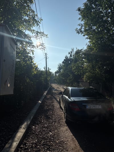 The image depicts a static outdoor scene on a bright, sunny day, likely in Bozhurishte, Bulgaria. The photograph is taken with the sun directly in front, resulting in significant lens flare and strong backlighting across the scene.
In the foreground and mid-ground, a dark-colored Mercedes-Benz sedan is parked on the right side of an unpaved road, which is covered with dark gravel or small stones. A concrete curb or barrier runs alongside the road, parallel to the car. The rear of the car is clearly visible, featuring a Bulgarian license plate that reads "CB 6466 KT" and the iconic Mercedes-Benz logo.
Lush green trees with dense foliage line both sides of the road, creating a verdant canopy that partially obscures the sky. On the left side of the frame, a wooden utility pole with multiple overhead wires is visible, positioned between the viewer and the bright sun. The sun is partially hidden by the foliage, casting strong rays and creating a clear blue sky above.
In the bottom-left foreground, a dark grey metal box or utility cabinet is partially visible, with some indiscernible text or stickers on its surface. The strong sunlight and clear sky suggest the time of day is likely mid-morning to early afternoon. There is no discernible activity taking place.