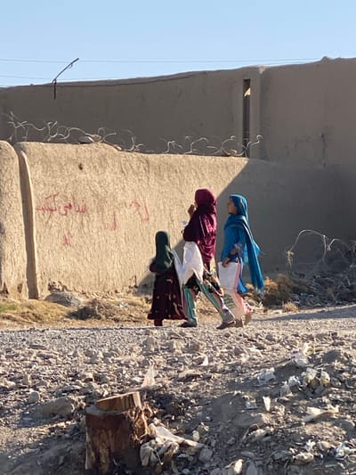 The image captures a ground-level view of a dusty, semi-urban environment in کوکران, Afghanistan. Three females are depicted walking from left to right on a rough, unpaved path composed of dirt, gravel, and scattered debris including plastic bags and other refuse. In the immediate foreground to the lower left, a dark brown tree stump is visible, surrounded by additional litter.
In the midground, the three females walk in single file. The leftmost figure, a younger female, wears a dark green head covering and a long, dark patterned dress. The middle figure, an adult female, wears a dark maroon head covering and a colorful, patterned long dress; she holds a folded white cloth or thin bag in her left hand and appears to be touching her face with her right hand. The rightmost figure, also an adult female, wears a vibrant blue head covering that drapes over her shoulders and a light-colored, patterned dress; she carries a white plastic bag with visible contents in her left hand. All three appear to be wearing open-toed footwear.
The background is dominated by a light-brown, textured mud or adobe wall, roughly constructed, stretching horizontally across the frame. Red Farsi script graffiti is visible on the lower-left section of this wall. Along the top edge of this wall, concertina barbed wire is installed. Further back, a darker, higher wall section or structure is visible, partially obscured by shadow. The sky above is clear and light blue, indicating daylight. Sunlight illuminates the scene from the right, casting shadows from the figures and structures onto the ground and the wall.
