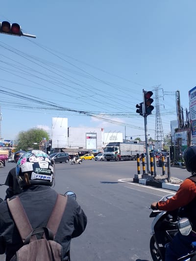 The image depicts a busy intersection in the city of Ciperna, Indonesia, under a clear blue sky. A traffic light is visible, indicating red, yellow, and green signals. Several vehicles, including motorcycles, cars, and a large truck, are navigating the intersection. Two individuals on motorcycles are in the foreground, one wearing a helmet adorned with stickers. A truck is in the process of turning, while other vehicles are stopped or moving. Power lines crisscross the sky above the intersection. Billboards are visible in the background, suggesting a commercial area. The overall scene captures a typical urban traffic scenario.