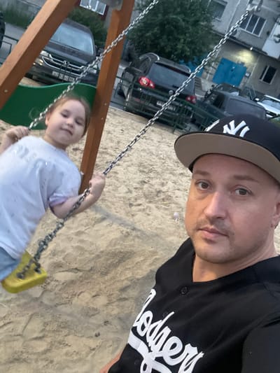 A man wearing a black baseball cap with a white "NY" logo and a black shirt with white lettering that reads "Dodgers" is taking a selfie. He is looking towards the camera with a neutral expression. To his right, a young girl with blonde hair and a white t-shirt is swinging on a wooden swing set. Her eyes are closed, and she appears to be enjoying the swing. The background shows a sandy playground area, with a building and parked cars visible in the distance. The time of day appears to be late afternoon or early evening, as indicated by the soft lighting. The overall scene suggests a casual, family-friendly outing in a residential area, possibly a playground in Surgut, Russia.