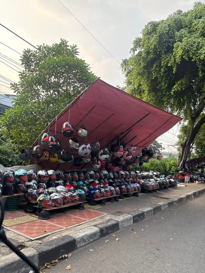 This media file captures an outdoor stall selling a large collection of motorcycle helmets. The scene is set on a sidewalk next to a paved road, with lush green trees and foliage providing a backdrop. A large, reddish-brown tarp is stretched over a simple frame, creating a shaded area for the display.
The primary objects are hundreds of motorcycle helmets, neatly arranged on racks and shelves. Some helmets are hung from the tarp's frame, while others are stacked in rows on tables and on the ground. They come in a variety of colors, designs, and types, including full-face helmets, open-face helmets, and what appear to be children's helmets. The sheer volume suggests a retail or rental operation.
The setting appears to be a street in South Jakarta, Indonesia, indicated by the presence of utility lines and the overall urban environment. The time of day seems to be late afternoon or early evening, as suggested by the quality of light, which casts long shadows and has a warm hue. The sky is partly cloudy. No people are clearly visible in the foreground, though some figures might be obscured by the helmets or out of frame. There is no visible text in the image. The overall impression is a bustling marketplace or a roadside vendor catering to motorcycle riders.