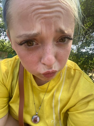A close-up, low-angle selfie shows a young person with fair skin, freckles, and bright green eyes looking directly at the camera with pursed lips and furrowed brows. They have long, dark false eyelashes and light brown hair with streaks of blue visible at the top left. The person is wearing a bright yellow t-shirt with thin white stripes. A brown strap is visible across their shoulder, and a silver necklace with a round pendant featuring a cat image hangs down. The background is slightly blurred, showing green trees and foliage, suggesting an outdoor setting, possibly a park or yard. There is no indication of the city of Nova Praha, Ukraine.