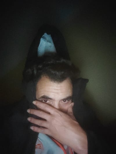 A person in a dark hoodie covers half their face with a hand, focusing intensely. The person's brown eyes are the most visible feature, contrasting with the shadowed setting. The hoodie's interior has a light blue or white lining. The background is indistinct, appearing as a dark, muted green or gray. The overall mood is enigmatic and slightly unsettling.
