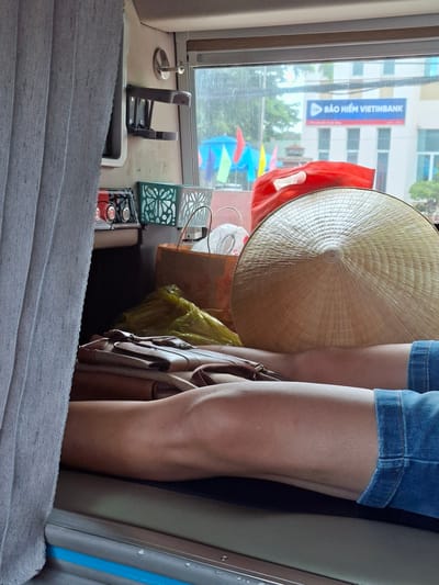 The image shows a person lying down inside what appears to be a sleeper bus. The person's legs, clad in denim shorts, are visible, resting on a surface with a brown bag and a conical hat nearby. The background reveals a window with a view of buildings and trees, suggesting an urban or semi-urban environment. A blue sign with Vietnamese text, "BẢO HIỂM VIETINBANK," is prominently displayed on one of the buildings, indicating the presence of a Vietinbank insurance branch. The overall scene is dimly lit, with the outside environment appearing overcast. The location is identified as Liên Nghĩa, Vietnam.