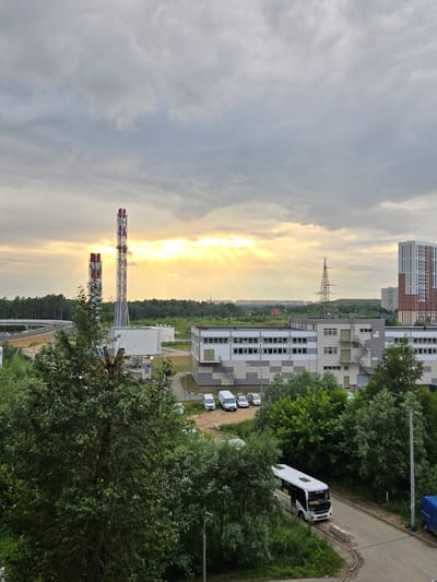 This is a medium-distance, high-angle exterior shot of an industrial or commercial area in Balashikha, Russia, under a cloudy sky with the sun breaking through. In the foreground, lush green trees dominate the left side of the frame, with a prominent tree trunk visible. To the right, a paved road curves through the scene, with a white bus parked on it. Further back, a long, multi-story building with a gray and white checkered facade features numerous windows and external staircases. Beyond this building, a tall lattice tower with power lines stands against the horizon. Two large industrial chimneys with red and white tops are visible on the left side of the frame, with a network of roads and a bridge structure behind them. In the far distance, a high-rise residential building with a distinctive red and white pattern is situated, along with other buildings and a dense line of trees marking the edge of the developed area. The sky is heavily clouded, with shafts of sunlight illuminating the scene from the left, creating a dramatic effect.