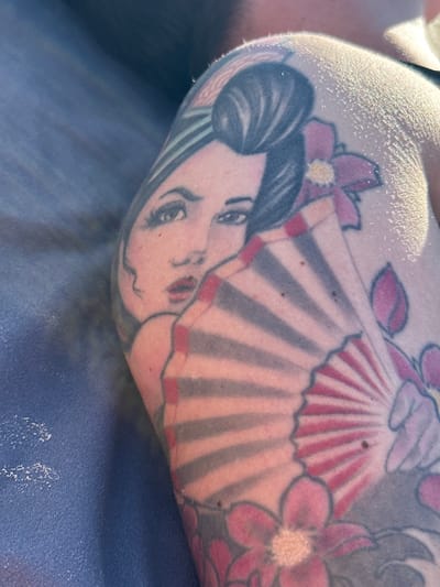 The image shows a close-up of a tattoo on a person's arm. The tattoo features a Japanese geisha with a fan and flowers. The skin appears to be lightly tanned and has some small water droplets or sweat. The background is blurred, but suggests an outdoor setting with blue and grey tones. There are no identifiable landmarks or context to determine a specific location in Jūrmala, Latvia.