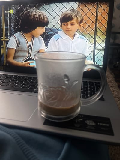 The image shows a laptop screen displaying a scene with two boys standing behind a diamond-patterned fence. The boys are wearing light-colored tops with trim. A partially filled glass mug is resting on the laptop keyboard. The laptop is gray, and a portion of a dark blue garment is visible on the right side of the frame. The TV channel "bTV 2" is visible on the lower left of the screen. The location in the city of Blaskovo, Bulgaria cannot be determined from this image.