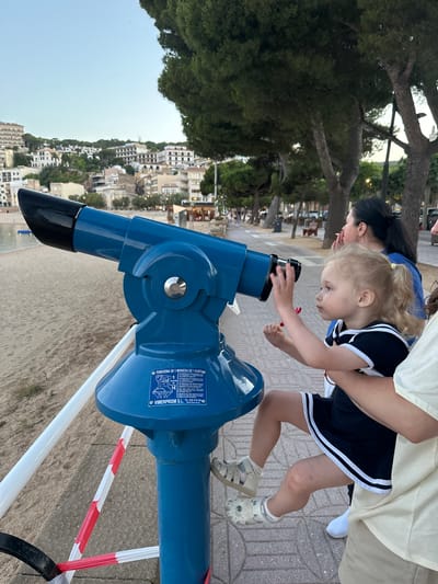 A young girl with blonde pigtails, wearing a black and white striped dress and white sandals, is looking through a large blue coin-operated telescope on a beach promenade. She is being held by an adult whose arm is visible. In the background, a woman with dark hair is visible looking towards the sea. The promenade features a paved walkway and a sandy beach with the sea in the distance. Buildings line the hillside behind the beach, with trees and streetlights visible along the promenade. The sky is clear and a light blue, indicating it is either early morning or late afternoon. Signage on the telescope indicates it requires one euro to operate and provides instructions in multiple languages. The overall scene suggests a recreational outing at a coastal tourist destination. The city of Sant Feliu de Guíxols, Spain is implied by the signage and the architectural style of the buildings, consistent with coastal towns in this region.