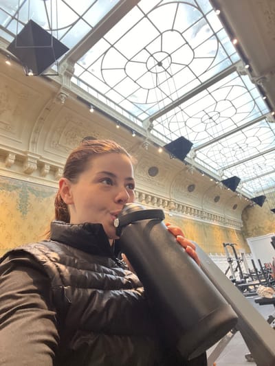A female individual is depicted in an indoor setting, observed drinking from a large, black insulated bottle. She wears a black puffer vest over a long-sleeved black garment. Her hair is pulled back, and her facial expression indicates she is actively consuming liquid from the bottle, which she holds with her right hand. The background features a distinctive, ornate architectural environment consistent with the gymnasium facilities located within La Piscine - Musée d'Art et d'Industrie André Diligent in Roubaix, France. Above the subject, a large, multi-paneled glass skylight ceiling with intricate circular and rectangular patterns provides natural illumination. The walls display classical moldings, cornices, and decorative friezes in a light tone, with a faded patterned section below. Several black, geometric acoustic panels or decorative elements are suspended from the ceiling beams. To the right of the subject, various pieces of gym equipment, including weight benches, are visible, confirming the fitness facility context.