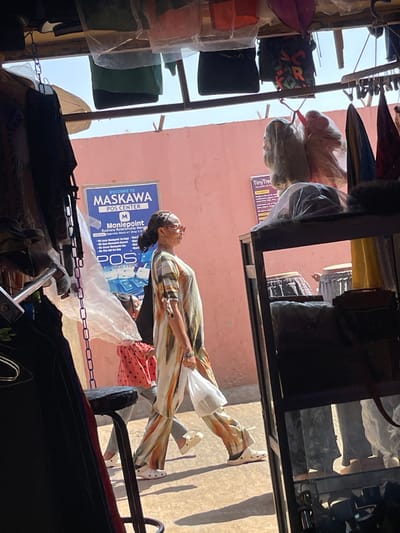 An outdoor scene in Samaru, Nigeria, is depicted from the interior of a commercial establishment. A woman walks from left to right across the midground, illuminated by strong sunlight, casting distinct shadows. She wears a long, loose-fitting, earth-toned patterned tunic and matching wide-leg trousers, white Crocs, and sunglasses, with her dark hair pulled back. She carries a white plastic bag in her left hand. Partially obscured behind her to the left is a child wearing a red and white polka-dot top and light-colored pants, also walking in the same direction.
The background features a plain pink-peach colored building wall. Affixed to this wall is a blue and white sign reading "WELCOME TO MASKAWA POS CENTER Moniepoint Business Relationship Manager" with a partial address "Opposite Block A1 Shop 5". Another smaller sign, partially visible to the right, displays "TinyTree". An outdoor canopy or umbrella is visible against the clear sky above the wall.
The foreground and framing elements indicate the viewpoint is from inside a shop. Hanging items, including dark garments and items in plastic bags, are visible along a beam across the top. A translucent white plastic sheet hangs on the left. A dark metal display rack containing various packaged goods, including what appear to be wigs or hair extensions in clear plastic bags, occupies the right side of the frame. A dark metal stool or chair is partially visible in the lower-left foreground.