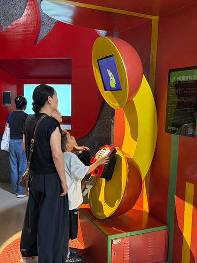 In the city of Jiangbei City Street, China, a simulated fire call exhibit is on display. The exhibit features a large, stylized red and yellow telephone receiver that is cut in half and appears to be an interactive screen. A woman, her back to the camera, stands with two children. One child, a boy, is actively engaged with a red and gray phone handset at the base of the exhibit, his hand on the keypad. The other child, also a boy, stands between the woman and the interactive phone, looking up at the screen. In the background, another woman wearing a blue shirt and carrying a white bag walks away from the exhibit. The environment is brightly colored, with red walls and yellow and green accents, suggesting an educational or recreational space, possibly a museum or exhibition hall. A sign with Chinese characters and "Fire Call 119 Simulation" in English is visible on the right side of the image.