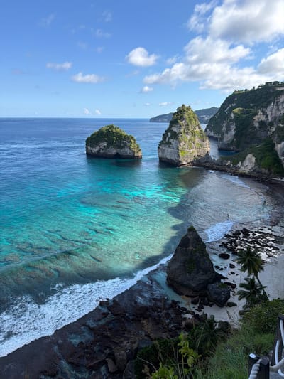 The image depicts an aerial view of the coastline in Pejukutan, Indonesia. The turquoise waters are dotted with several rocky islets covered in vegetation. The sea transitions to a lighter, more transparent hue near the shoreline, revealing coral reefs beneath the surface. White waves crash against the black rocky beach. Some sandy patches with palm trees are present near the coast. The sky is blue with scattered white clouds. Some ropes and vegetation are visible in the upper right corner, indicating a viewpoint.